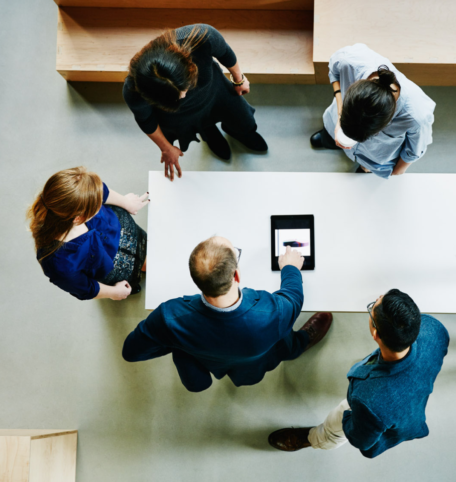 Aerial image of a group of professionals, standing around a desk, observing a tablet.